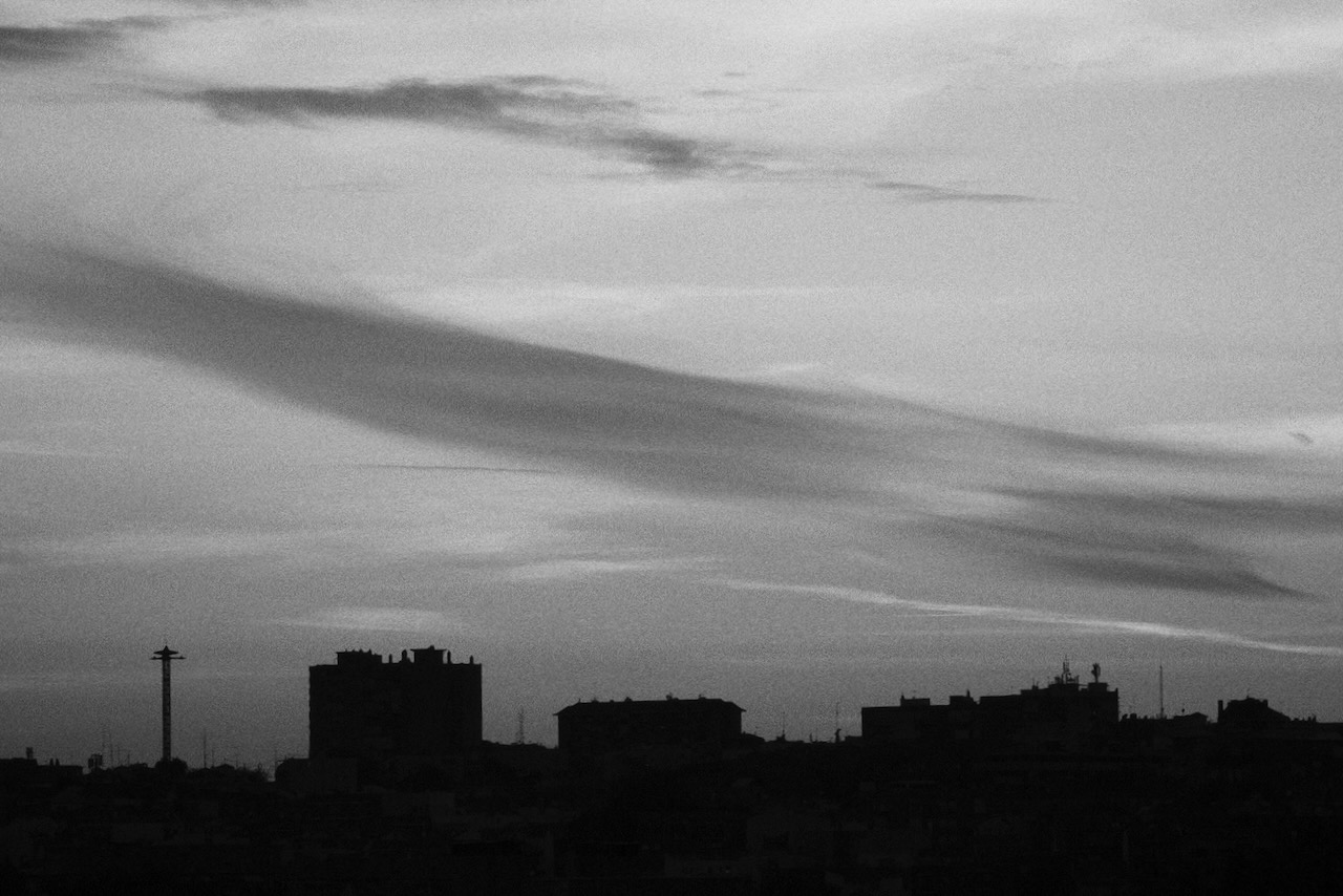 Black and white view of a city skyline in silhouette beneath a wide sky streaked with soft, wispy clouds at dusk.