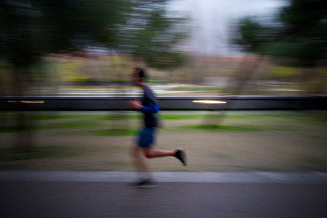 A blurred man running on a paved path near a fountain and trees.