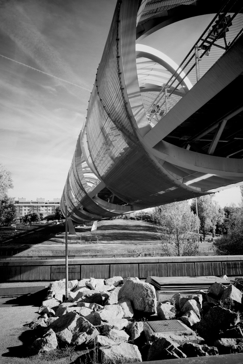 A modern, curved bridge with metal railings over a river, surrounded by rocks and trees.