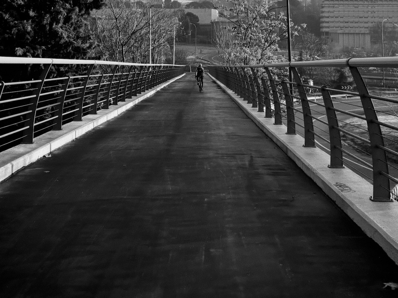 A cyclist on a wet, elevated pathway with metal railings, surrounded by trees and urban structures.