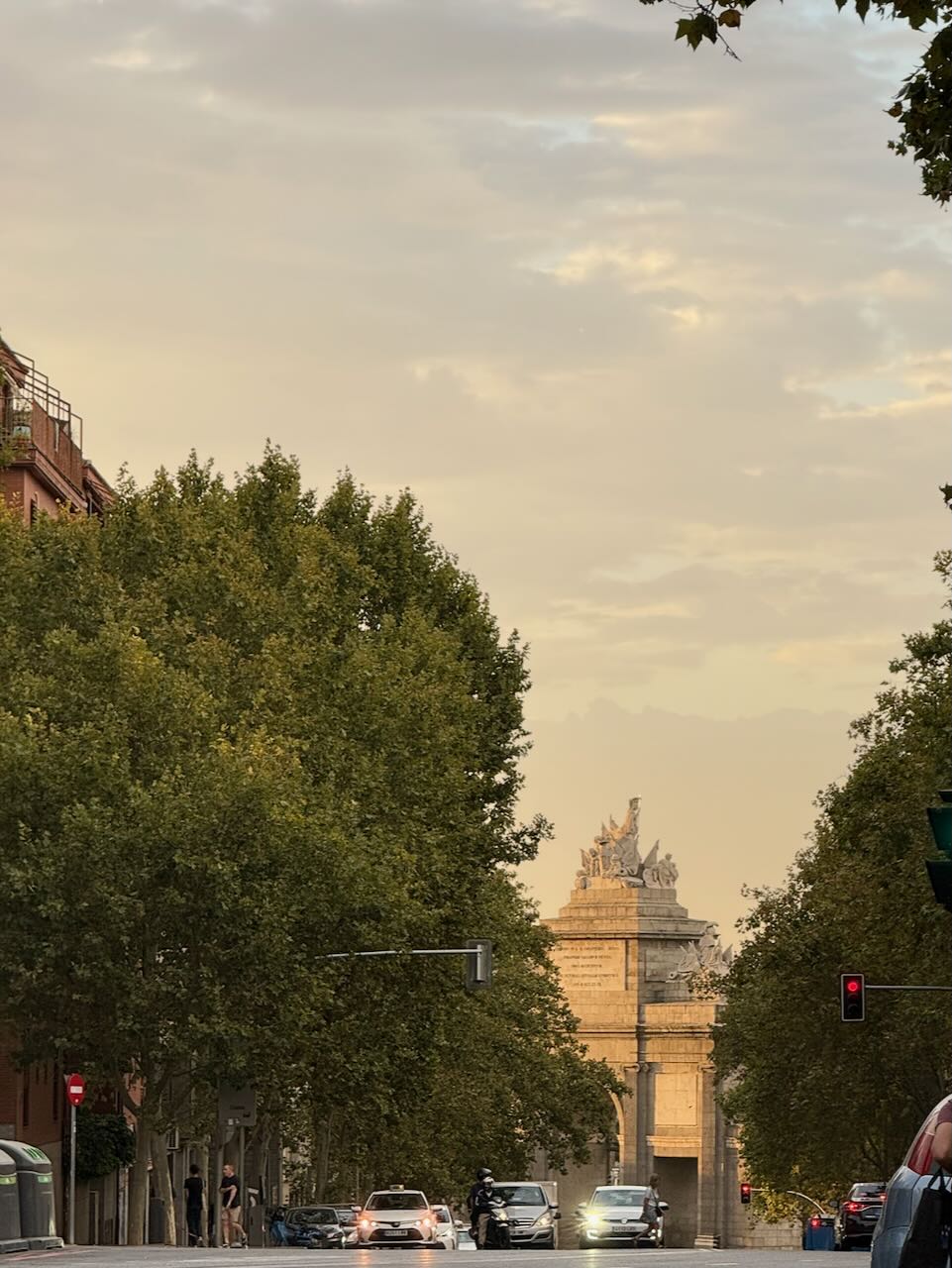 A city street with trees, cars, and a stone archway under a cloudy sky.